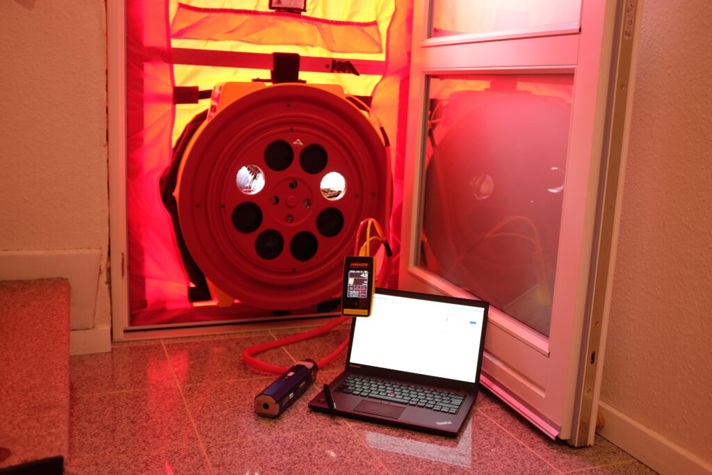 Test szczelności domu: wentylator blower door w czerwonej ramie z tkaniny zamontowany w otworze drzwiowym, obok na posadzce laptop i miernik ciśnienia