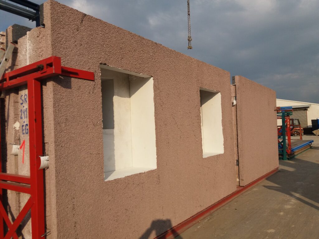 Prefabrykowane ściany z keramzytobetonu z otworami okiennymi stoją w stalowych stojakach na placu producenta, nad nimi zwisa hak dźwigu