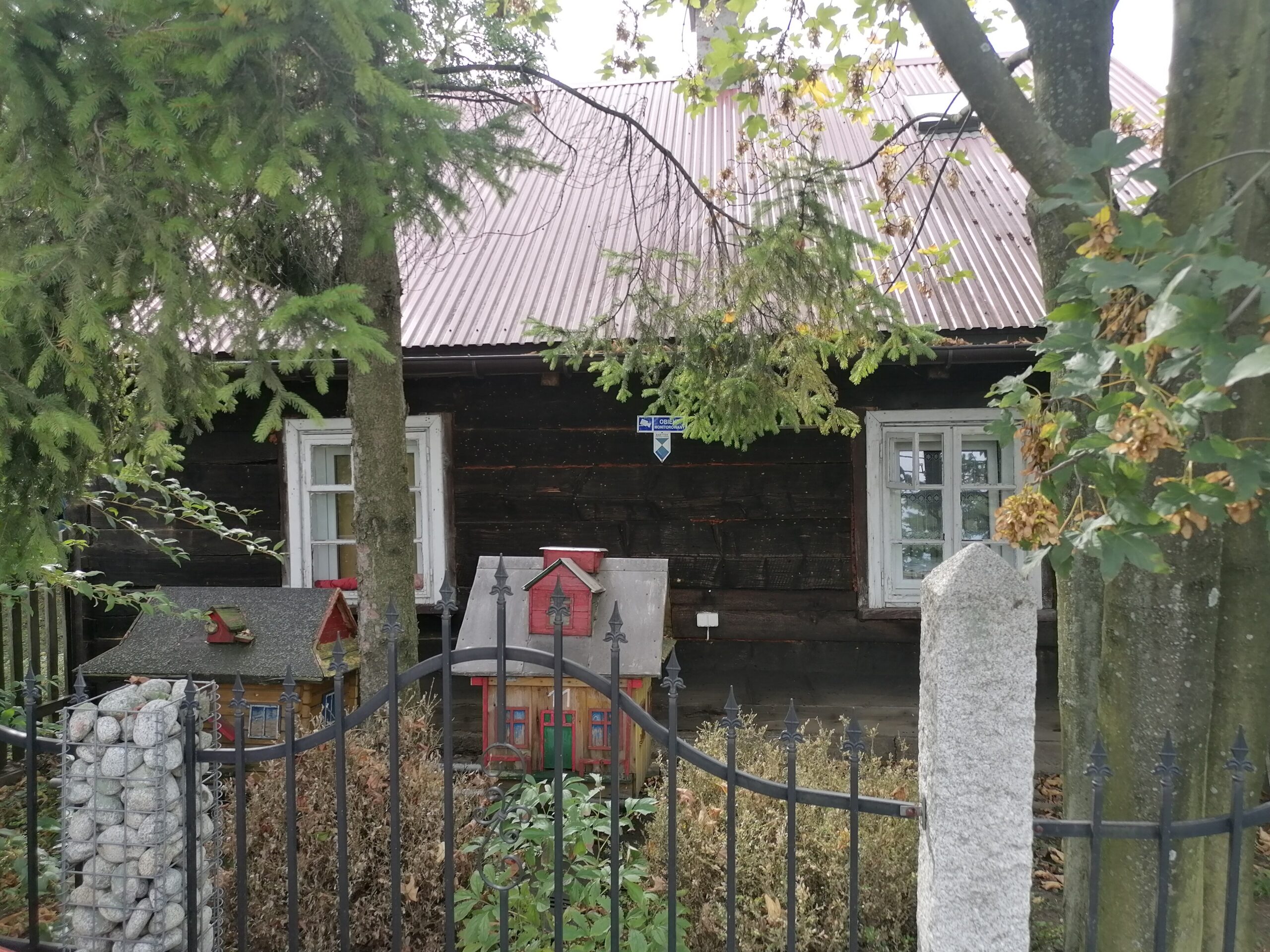 Stary dom z ciemnych bali z dachem z blachy i białymi oknami, za metalowym ogrodzeniem i drzewami