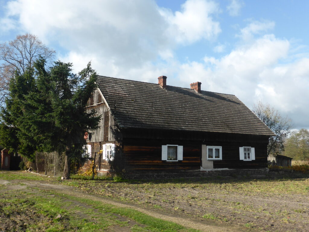 Stary parterowy dom z bali z dachem krytym dachówką, na niskiej kamiennej podmurówce, z deskowanym szczytem i białymi okiennicami