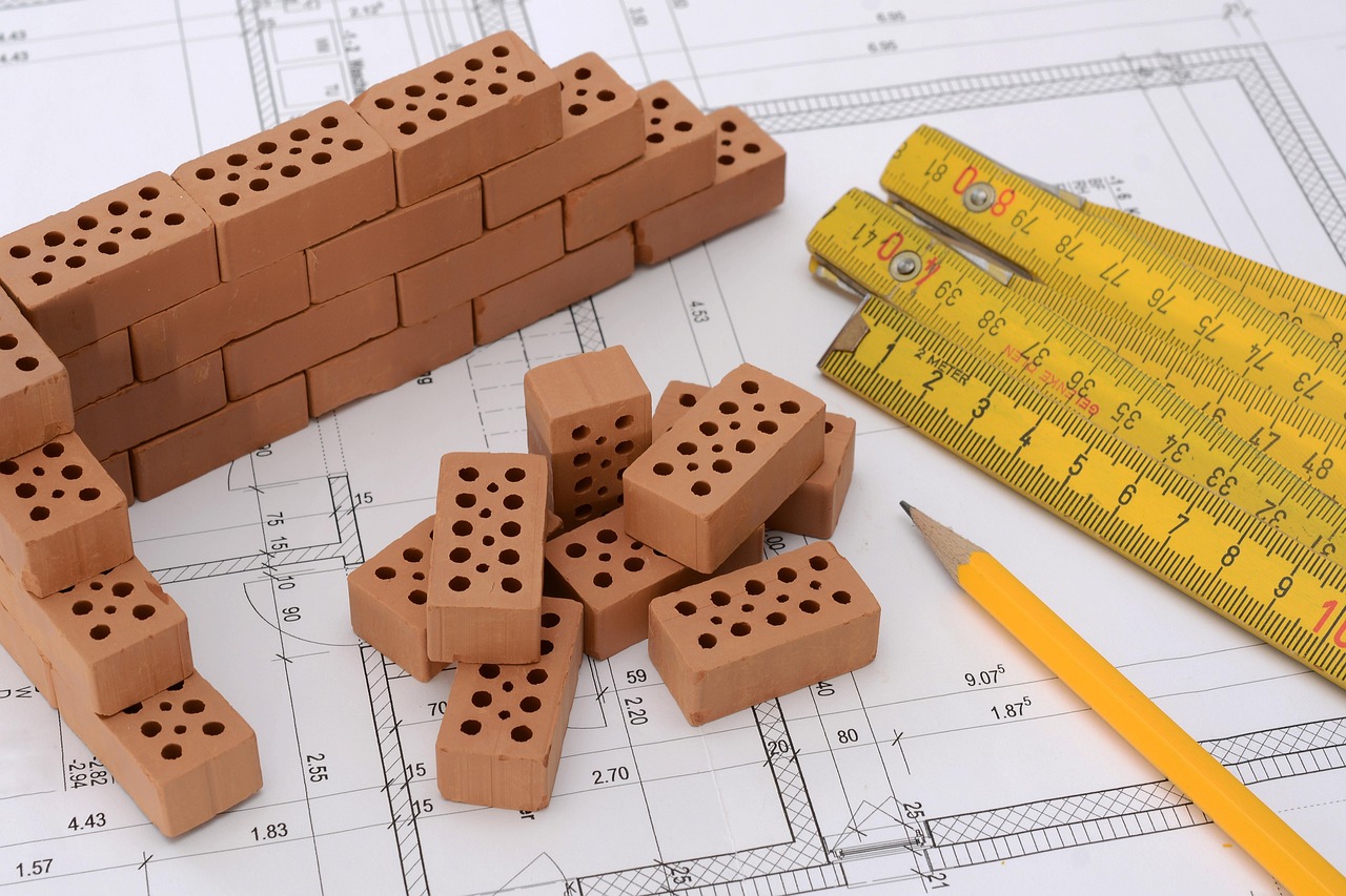 Rzut projektu domu z wymiarami, a na nim miniaturowe cegły, składana miarka i ołówek